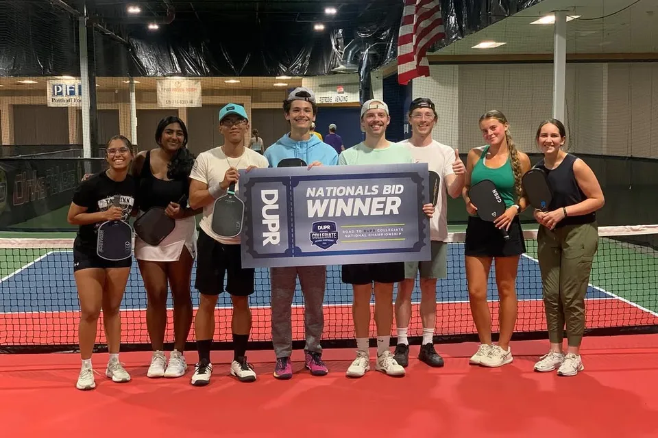 Nine members of a college pickleball team standing on an indoor court, holding a large DUPR 'Nationals Bid Winner' sign.